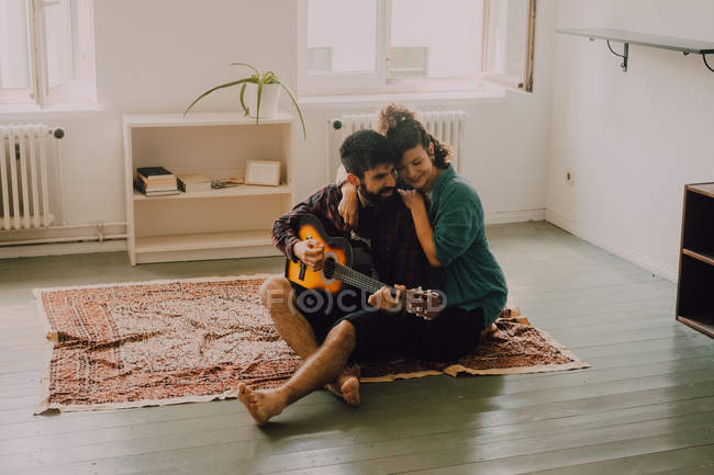 Tierna pareja alegre en trajes casuales tocando la guitarra en casa - foto de stock