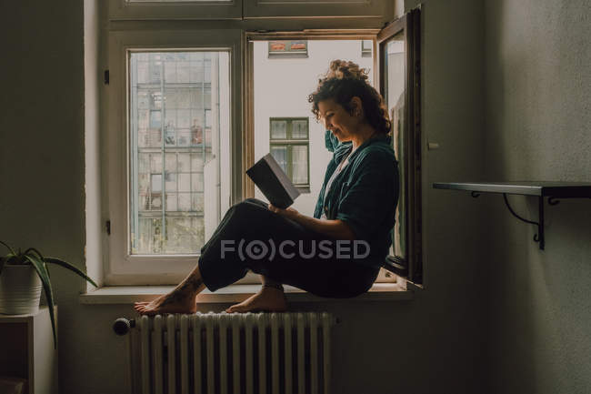 Seitenansicht einer entspannten Frau in lässigem Outfit, die Buch liest, während sie barfuß auf der Fensterbank in der Wohnung sitzt — Stockfoto