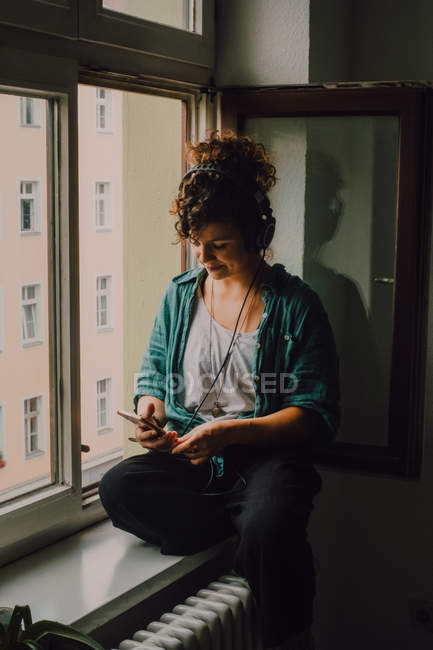 Усміхнена кучерява жінка в навушниках слухає музику під час перегляду смартфона і сидить на підвіконні в квартирі — стокове фото