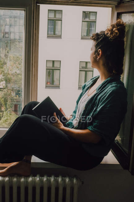 Side view of relaxed woman in casual outfit holding a book while sitting barefoot on window sill in apartment looking out of the window — Stock Photo