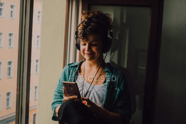 Smiling curly woman in headphones listening to music while browsing smartphone and sitting on window sill in apartment — Stock Photo