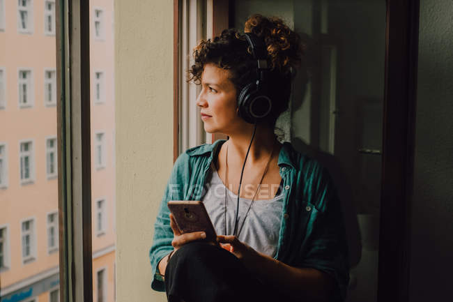 Pensive curly woman in headphones listening to music while browsing smartphone and sitting on window sill in apartment looking out of the window — Stock Photo