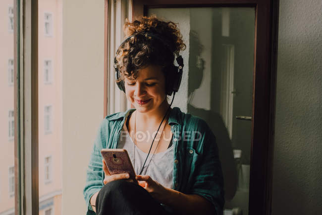 Mulher encaracolado sorridente em fones de ouvido ouvir música enquanto navega smartphone e sentado no peitoril da janela no apartamento — Fotografia de Stock