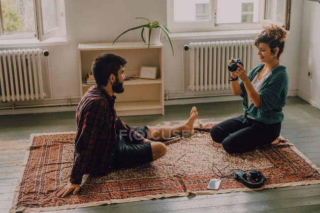 Side view of hipster woman taking picture of man sitting barefoot and posing on apartment floor — Stock Photo