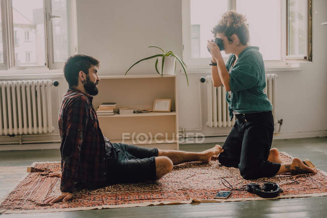 Vista laterale della donna hipster che scatta foto dell'uomo seduto a piedi nudi e posa sul pavimento dell'appartamento — Foto stock