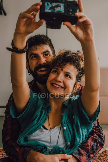 Affectionate hipster couple hugging and taking selfie on camera while sitting on floor at home — Stock Photo