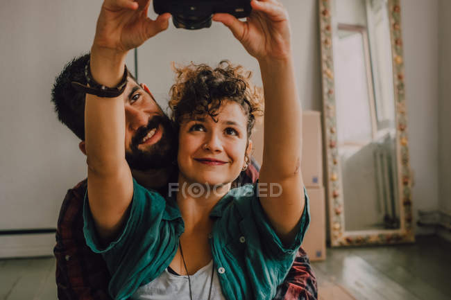 Ласковая пара хипстеров обнимает и делает селфи с камерой, сидя на полу дома — стоковое фото