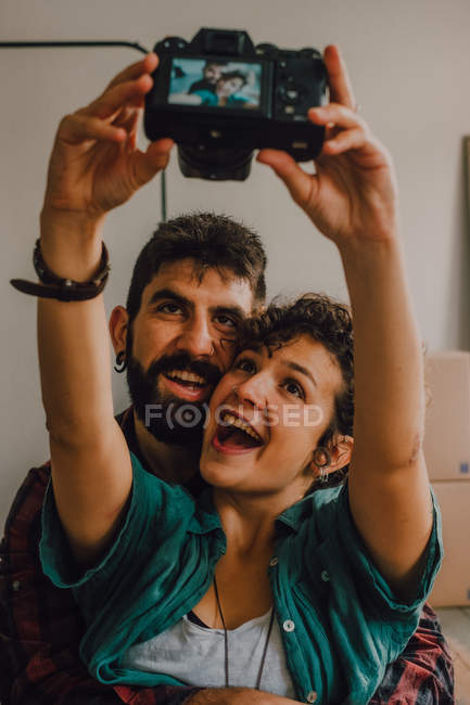 Casal hipster afetuoso abraçando e tirando selfie com câmera enquanto sentado no chão em casa — Fotografia de Stock