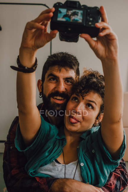 Affectionate hipster couple hugging and taking selfie with camera while sitting on floor at home — Stock Photo