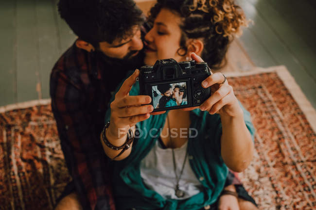 Blick von oben auf liebevolles Hipster-Paar, das sich küsst und Selfie mit Fotokamera macht, während es zu Hause auf dem Boden sitzt — Stockfoto