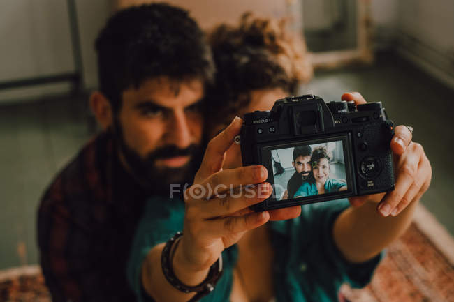 Desde arriba vista de pareja hipster cariñoso abrazo y tomar selfie con cámara de fotos mientras está sentado en el suelo en casa - foto de stock