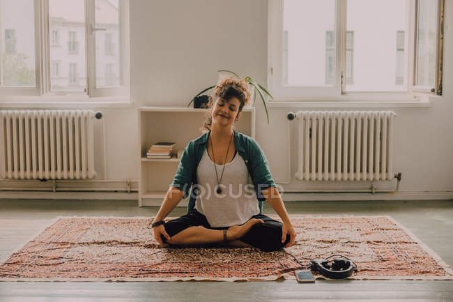 Mulher relaxada em roupas casuais sorrindo e sentada em lótus posar no tapete no apartamento moderno — Fotografia de Stock