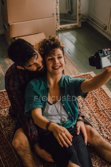 Сверху вид ласковой хипстерской пары, обнимающей и делающей селфи с фотокамерой, сидящей на полу дома — стоковое фото