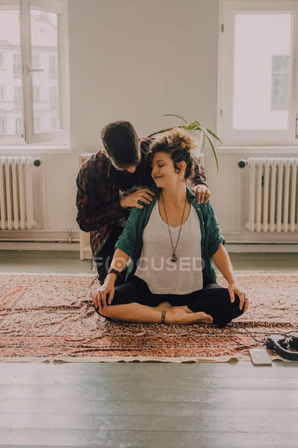 Hombre con ropa casual masajeando hombros de mujer relajada sentada en posición de loto en el suelo en casa - foto de stock