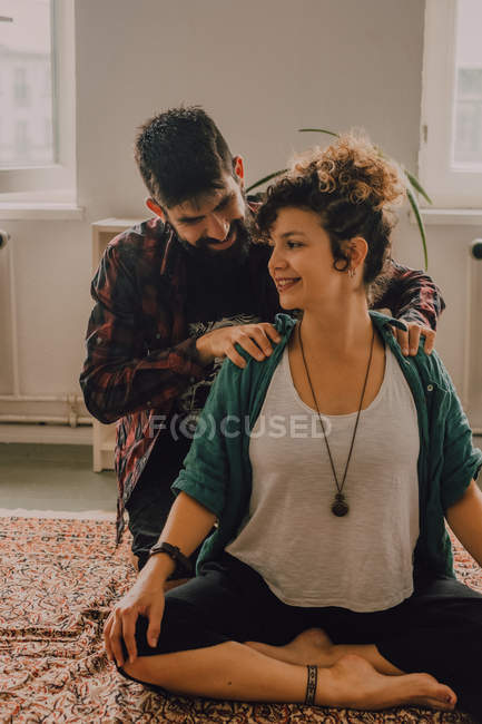 Man in casual clothing massaging shoulders of relaxed woman sitting in lotus position on floor at home — Stock Photo