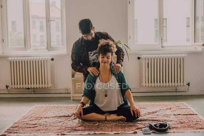 Homem em roupas casuais massageando ombros de mulher relaxada sentada na posição de lótus no chão em casa — Fotografia de Stock