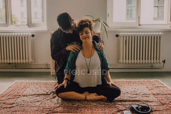 Homem em roupas casuais massageando ombros de mulher relaxada sentada na posição de lótus no chão em casa — Fotografia de Stock