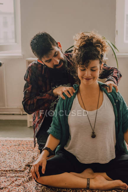 Мужчина в повседневной одежде массирует плечи расслабленной женщины, сидящей в позе лотоса на полу дома — стоковое фото