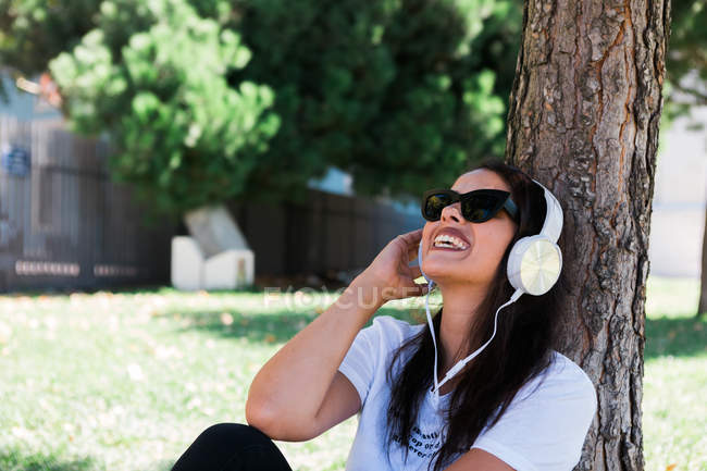 Мирная женщина в солнечных очках и наушниках слушает музыку, сидя на траве в парке — стоковое фото