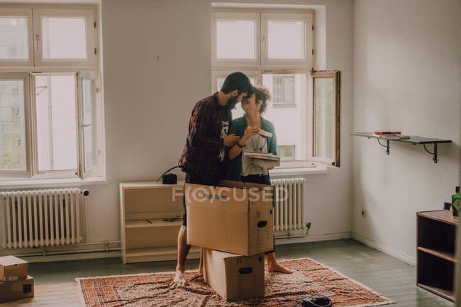 Пара хипстеров распаковывает вместе коробки, стоя босиком в светлой комнате и обнимаясь — стоковое фото