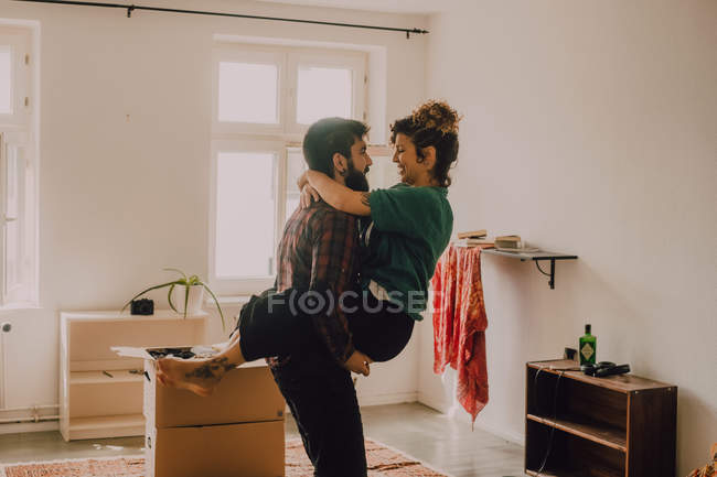 Вид сбоку влюбленной пары, обнимающейся и несущейся впереди, стоя рядом со стопкой картонных коробок дома — стоковое фото