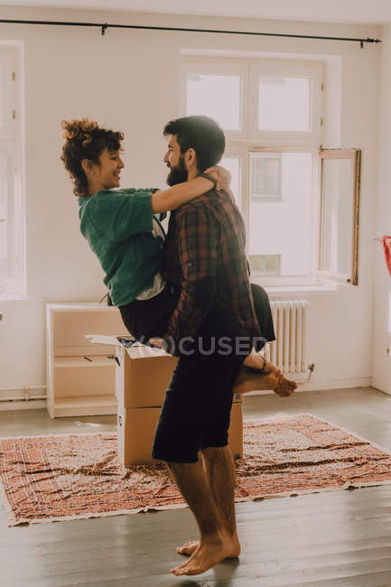 Side view of affectionate couple cuddling and front carrying while standing beside stack of cardboard boxes at home — Stock Photo
