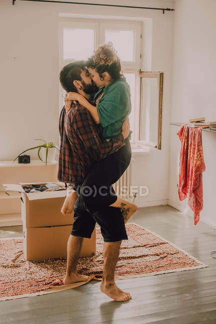 Vue latérale d'un couple affectueux câlinant et portant devant tout en s'embrassant à côté de la pile de boîtes en carton à la maison — Photo de stock