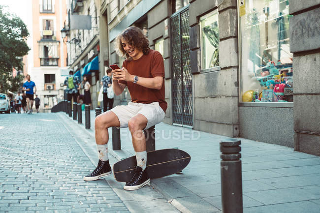 Alegre homem casual rindo e falando no smartphone enquanto sentado em balaústre na rua da cidade — Fotografia de Stock