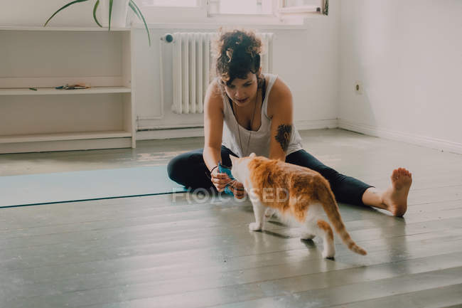 Cuidando de mulher casual dando comida para gato curioso enquanto sentado descalço em apartamento moderno minimalista — Fotografia de Stock