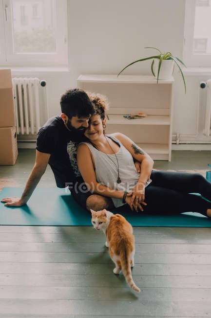 Vista laterale della coppia tenera che si abbraccia mentre si siede sul pavimento accanto al gatto rossiccio a casa — Foto stock