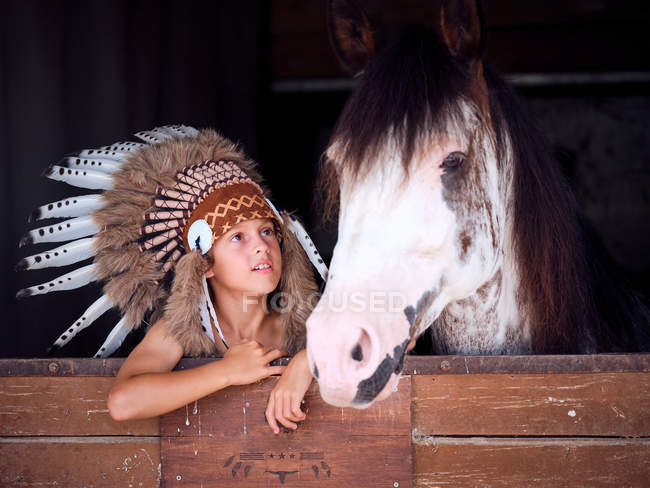 Очарованный ребенок в настоящей индийской шляпе из перьев опирается на деревянный забор в конюшне с лошадью на ранчо и смотрит в сторону — стоковое фото