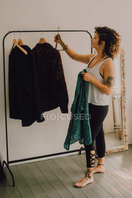 Vista lateral de la mujer casual colgando camisas en perchas mientras está de pie descalzo en la habitación minimalista - foto de stock