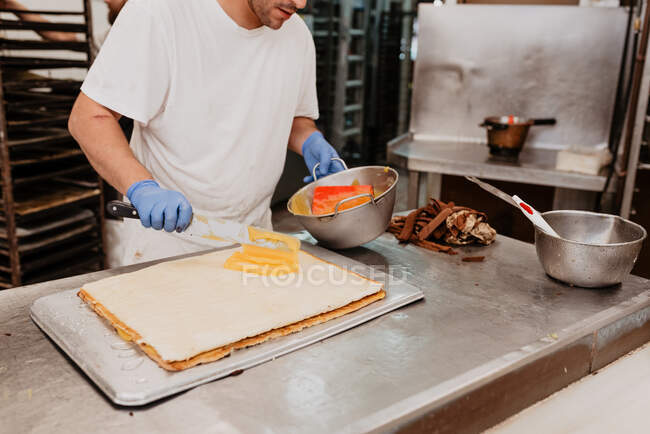 Bakery worker in latex gloves spreading sweet jam on fresh bun over kitchen counter — Stock Photo