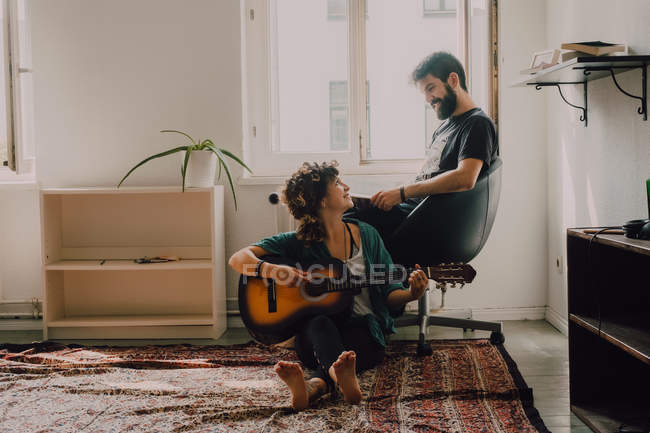 Mujer feliz tocando la guitarra y sentado en el suelo mientras el hombre lee el libro y escucha - foto de stock