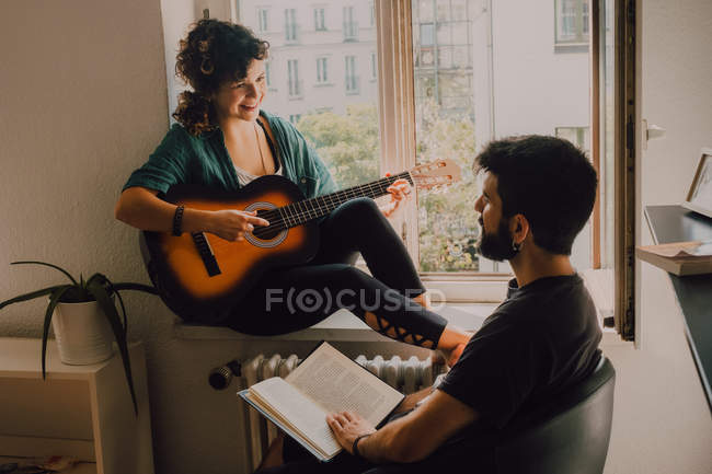 Mujer feliz tocando la guitarra y sentado en el alféizar de la ventana mientras el hombre leyendo libro y escuchando - foto de stock