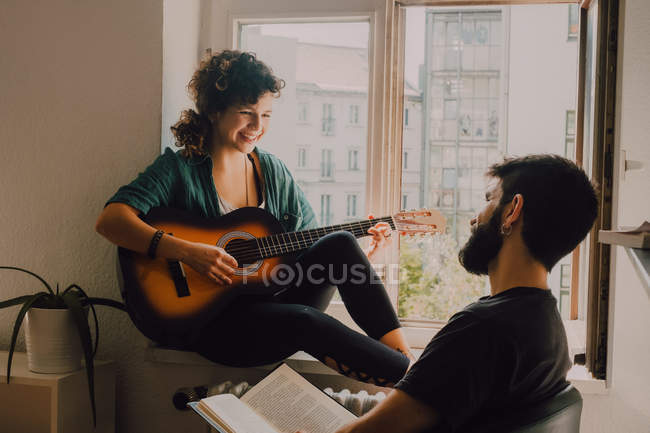 Счастливая женщина играет на гитаре и сидит на подоконнике, в то время как мужчина читает книгу и слушает — стоковое фото