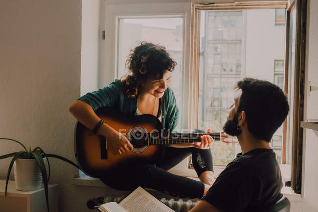 Щаслива жінка грає на гітарі і сидить на підвіконні, коли чоловік читає книгу і слухає — стокове фото