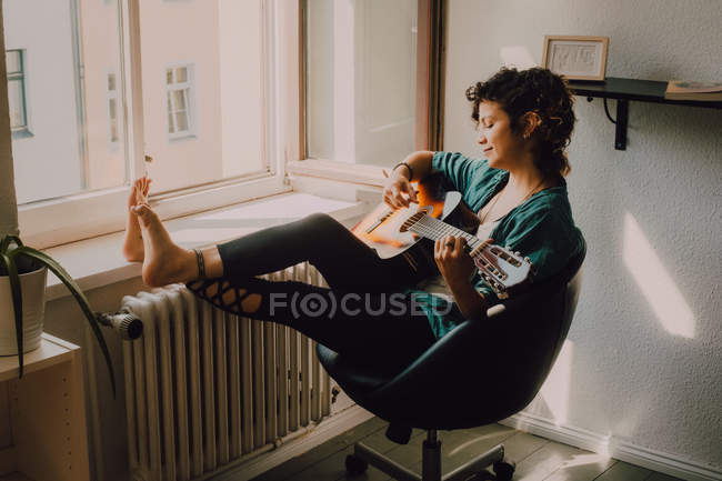 Femme pieds nus détendue en vêtements décontractés jouant de la guitare acoustique assis sur une chaise à côté de la fenêtre à la maison — Photo de stock