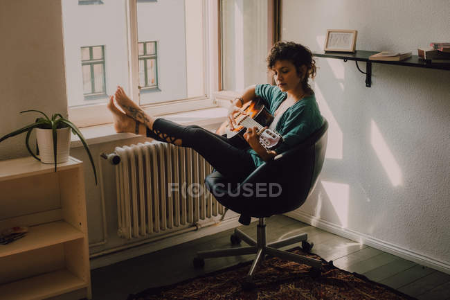 Femme pieds nus détendue en vêtements décontractés jouant de la guitare acoustique assis sur une chaise à côté de la fenêtre à la maison — Photo de stock