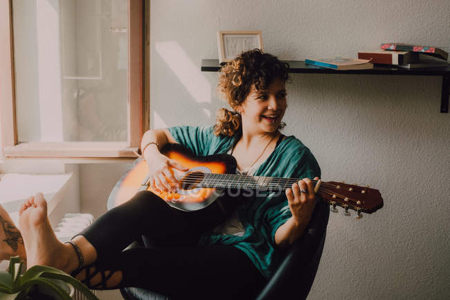 Entspannte barfüßige Frau in lässiger Kleidung spielt Akustikgitarre, während sie zu Hause auf einem Stuhl neben dem Fenster sitzt — Stockfoto