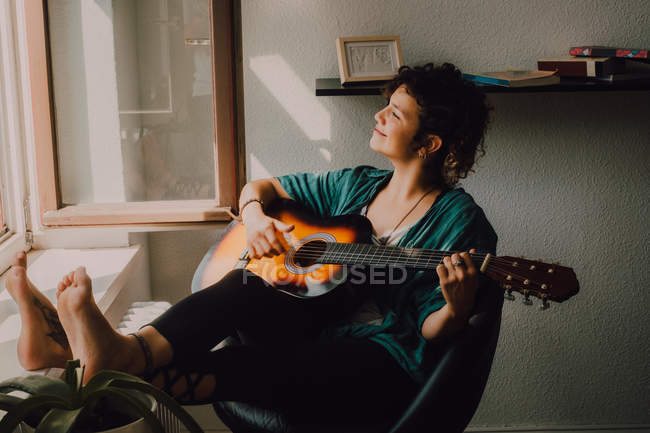 Расслабленная босиком женщина в повседневной одежде играет на акустической гитаре, сидя на стуле рядом с окном дома — стоковое фото