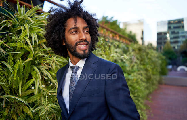 Optimistic african american beaded male entrepreneur in formal clothing looking to right side while standing near hedge — Stock Photo