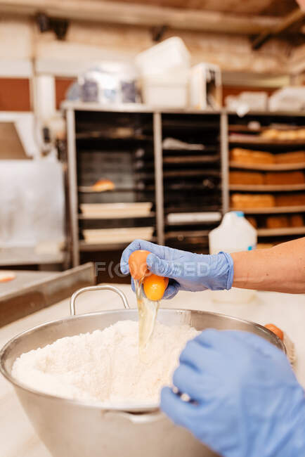 De cima confeiteiro irreconhecível em luvas de látex quebrando ovos de galinha frescos em tigela com farinha de trigo enquanto prepara pastelaria na cozinha — Fotografia de Stock
