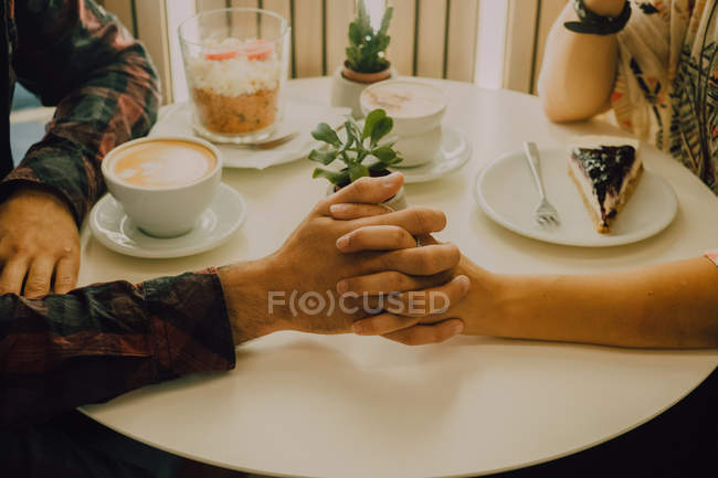 Vista de cerca de la pareja cariñosa cogida de la mano mientras está sentada en la mesa en la cafetería - foto de stock