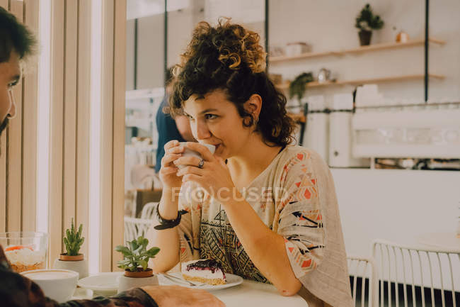 Vista lateral de la mujer sonriente feliz bebiendo de la taza mientras está sentado en la cafetería moderna - foto de stock