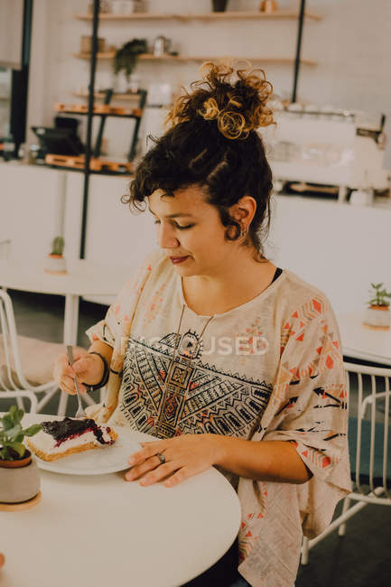 Радостная случайная женщина ест торт с вилкой, сидя за столом в кафетерии — стоковое фото