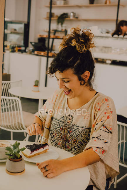 Радостная случайная женщина ест торт с вилкой, сидя за столом в кафетерии — стоковое фото