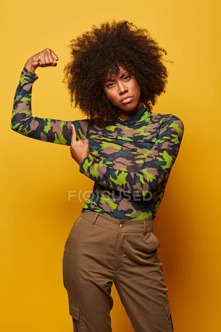 Serious strong African American woman in camouflage shirt showing biceps on yellow background looking at camera — Stock Photo