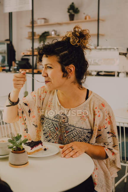 Joyful casual woman eating cake with fork while sitting at table in cafeteria — Stock Photo