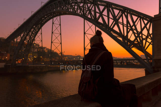 Back view of female tourist looking away while sitting on border of city embankment near bridge during sunset in Porto, Portugal — Stock Photo